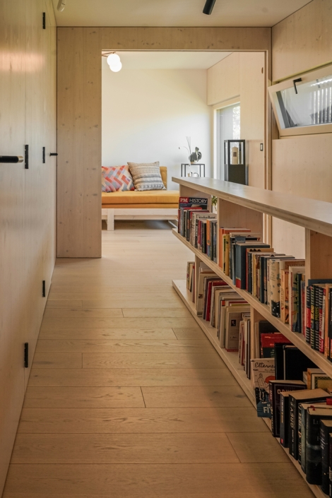 Hallway with a low bookshelf filled with books, leading to a cozy seating area with cushions.