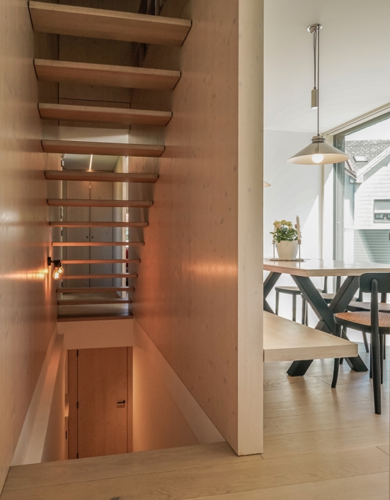 Wooden staircase above a hallway, next to a dining area with a table and potted plant.
