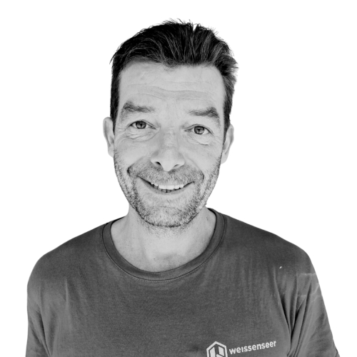 Porträtfoto eines Mannes in einem T-Shirt, der freundlich in die Kamera lächelt. Schwarz-weiß aufgenommen vor weißem Hintergrund.